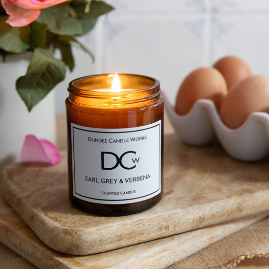 A lit amber glass scented candle jar with a white label reading 'Dundee Candle Works DCw Earl Grey & Verbena' sits on a wooden board in a kitchen setting, with flowers, eggs, and burlap visible in the background.