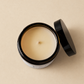 An open black glass jar candle with cream-colored wax and a single wick, shown with its black lid resting beside it on a warm beige background.