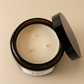 An open amber glass jar candle with a black lid leaning against it, showing creamy off-white wax with three wicks, on a warm beige background.