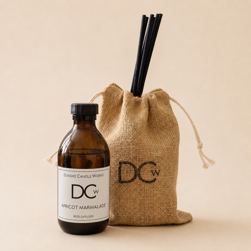An amber glass reed diffuser bottle with a black cap and white label next to a natural burlap drawstring bag with black reeds sticking out, on a warm beige background. The bottle label reads Dundee Candle Works DCw Apricot Marmalade Reed Diffuser.