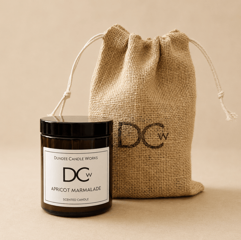 A scented candle jar with a black lid and white label in front of a natural burlap drawstring pouch. The label reads 'DUNDEE CANDLE WORKS DCw APRICOT MARMALADE SCENTED CANDLE'.