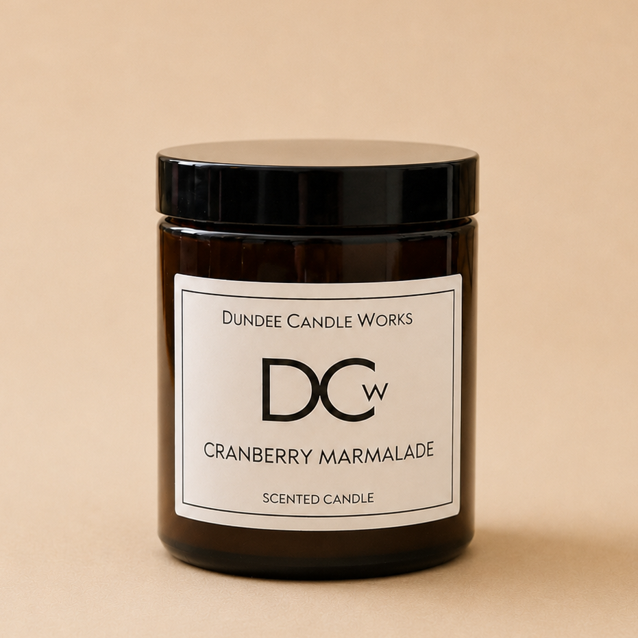 A dark amber glass jar candle with a black lid and a white label reading 'Dundee Candle Works DCW Cranberry Marmalade Scented Candle' on a warm beige background.