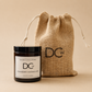A scented candle jar with a black lid and white label reading Dundee Candle Works DCw Cranberry Marmalade, placed in front of a natural burlap drawstring pouch with a large black DCw logo, on a warm beige background.