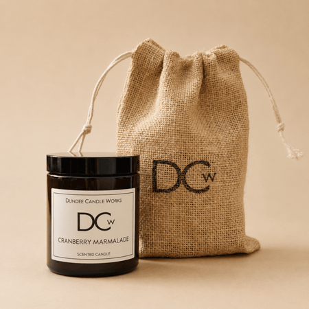 A scented candle jar with a black lid and white label reading Dundee Candle Works DCw Cranberry Marmalade, placed in front of a natural burlap drawstring pouch with a large black DCw logo, on a warm beige background.