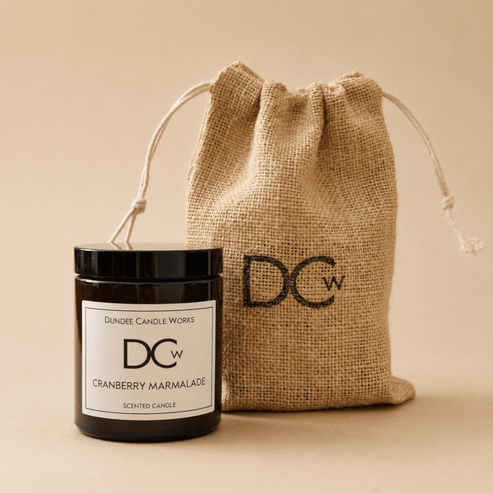 A scented candle jar with a black lid and white label reading Dundee Candle Works DCw Cranberry Marmalade, placed in front of a natural burlap drawstring pouch with a large black DCw logo, on a warm beige background.