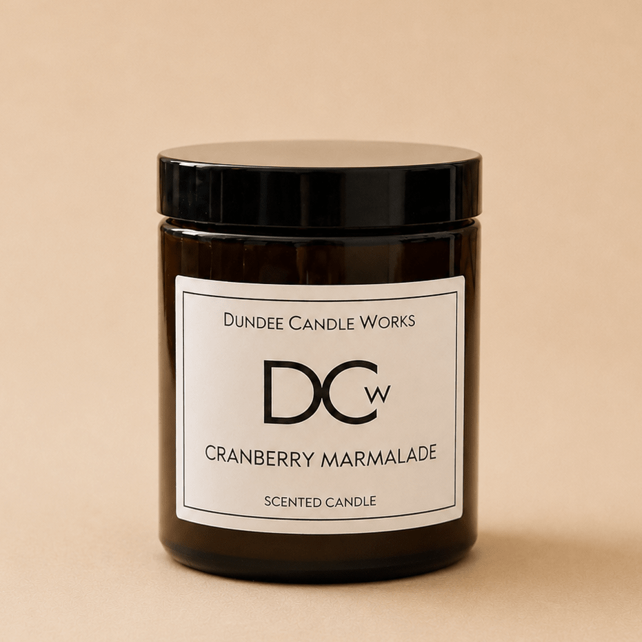 A dark amber glass jar candle with a black lid and a white label reading 'Dundee Candle Works DCW Cranberry Marmalade Scented Candle' on a warm beige background.