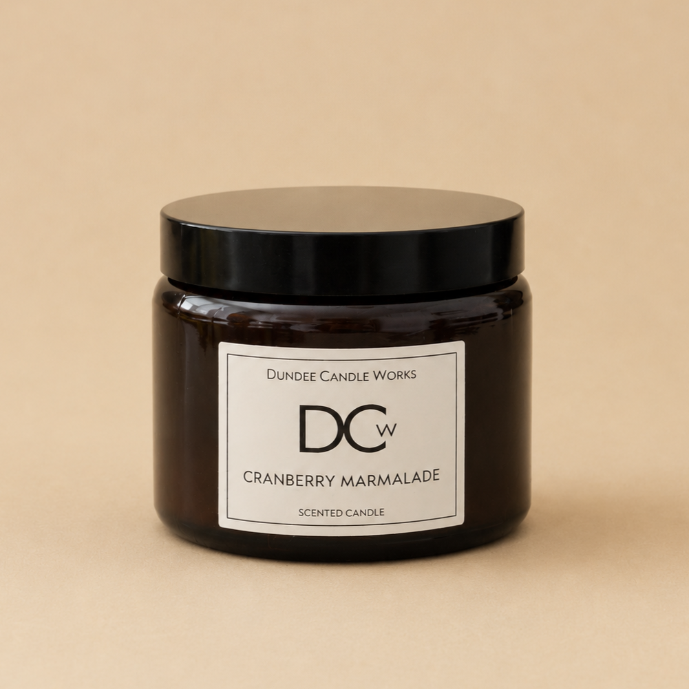 A dark amber glass jar candle with a black lid and a white label reading 'Dundee Candle Works DCW Cranberry Marmalade SCENTED CANDLE' on a warm beige background.
