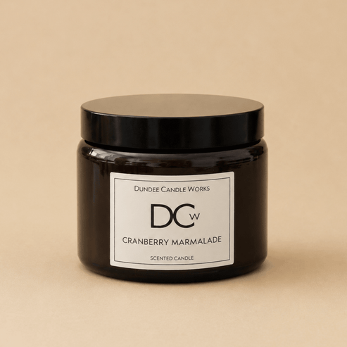A dark amber glass jar candle with a black lid and a white label reading 'Dundee Candle Works DCW Cranberry Marmalade SCENTED CANDLE' on a warm beige background.