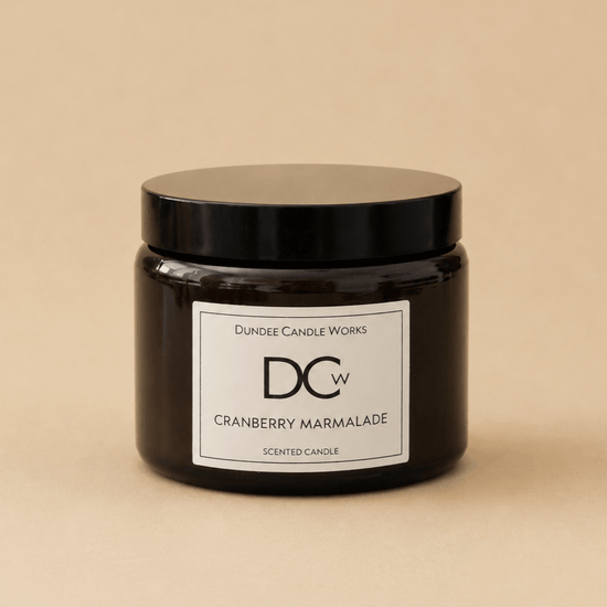 A dark amber glass jar candle with a black lid and a white label reading 'Dundee Candle Works DCW Cranberry Marmalade SCENTED CANDLE' on a warm beige background.