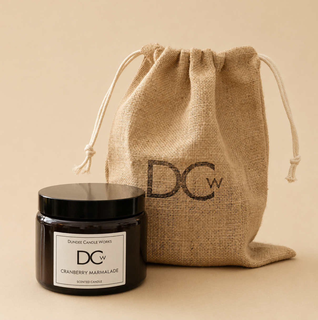 A dark glass candle jar with a black lid and a white label reading 'Dundee Candle Works DCw Cranberry Marmalade scented candle' placed in front of a natural jute drawstring pouch printed with 'DCW' on a warm beige background.