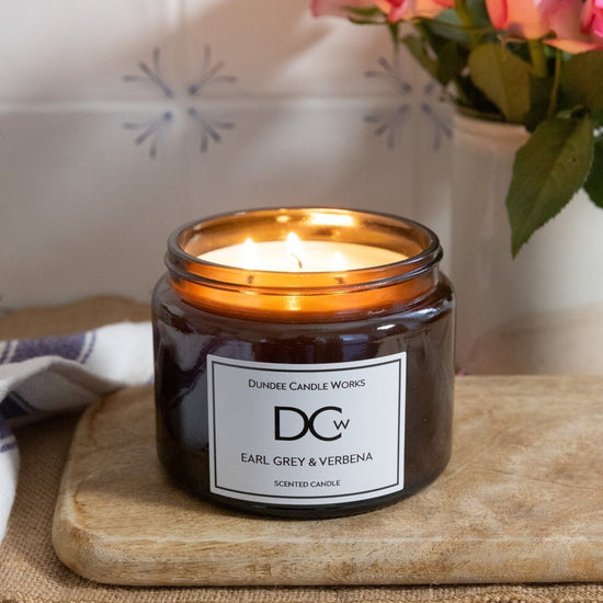 A lit scented candle in a dark amber glass jar sits on a wooden cutting board. The label reads 'Dundee Candle Works' and 'Earl Grey & Verbena'.