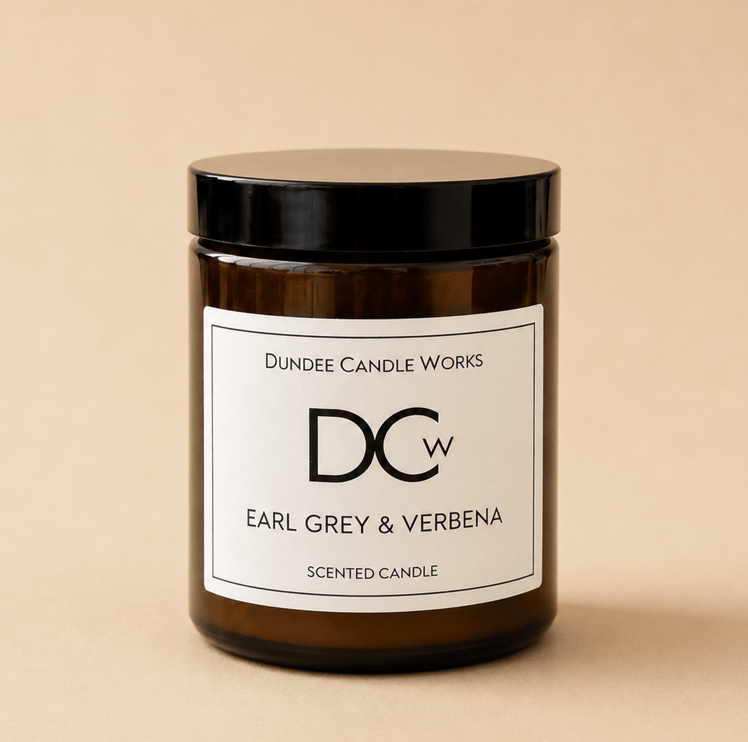 An amber glass jar candle with a black lid and a white label reading 'Dundee Candle Works DCW Earl Grey & Verbena Scented Candle' on a warm beige background.