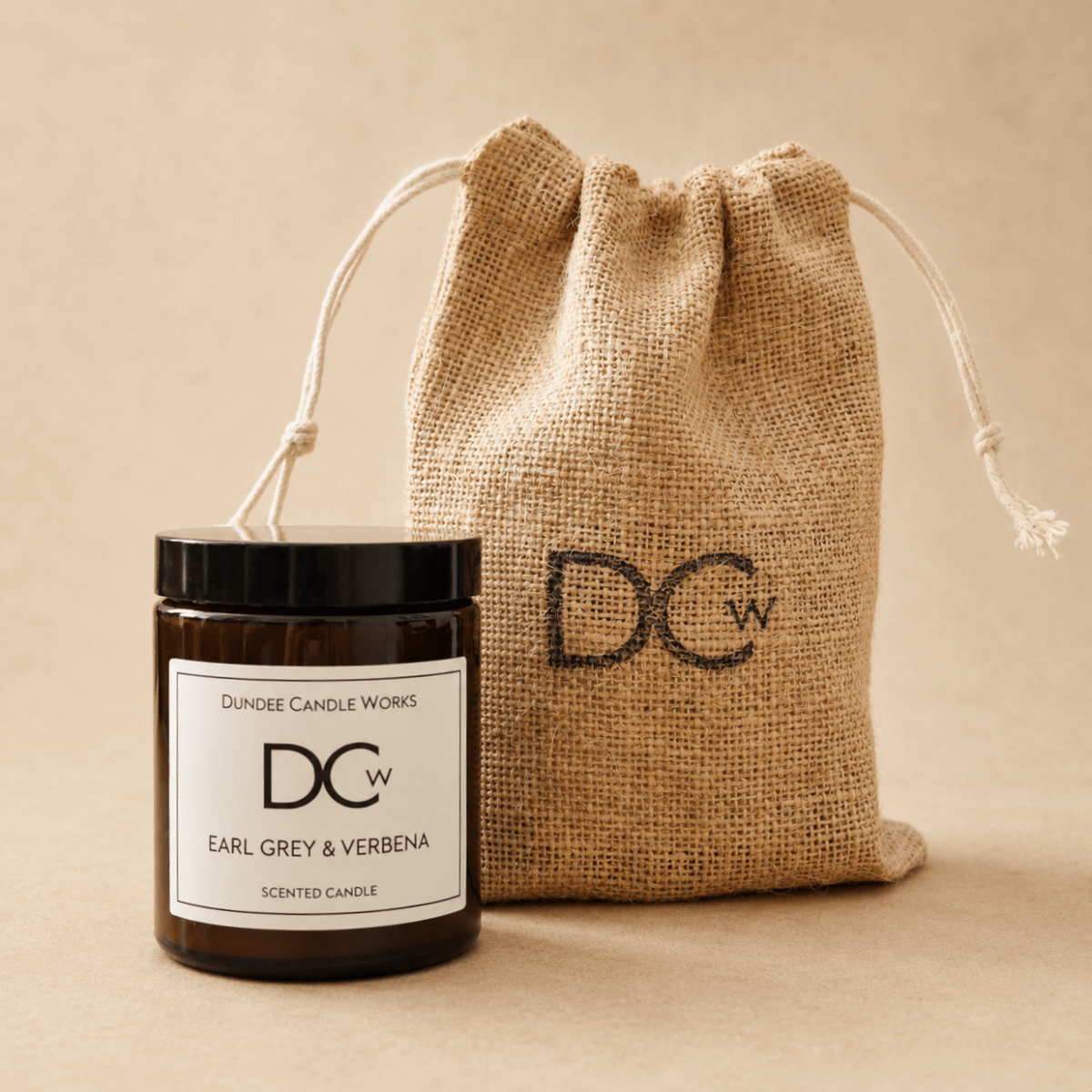 An Earl Grey & Verbena candle in a dark glass jar with a black lid and white label sits in front of a natural jute drawstring pouch. The pouch is printed with the brand initials 'DCw'.