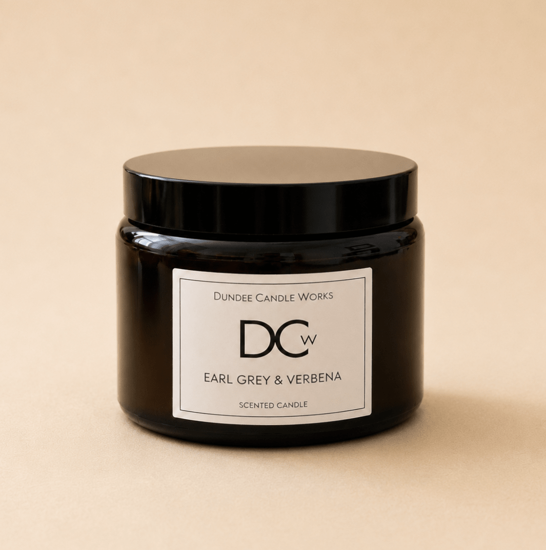 A dark amber glass jar candle with a black lid and a white label reading 'Dundee Candle Works DCw Earl Grey & Verbena Scented Candle' on a warm beige background.