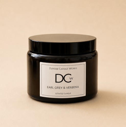A dark amber glass jar candle with a black lid and a white label reading 'Dundee Candle Works DCw Earl Grey & Verbena Scented Candle' on a warm beige background.