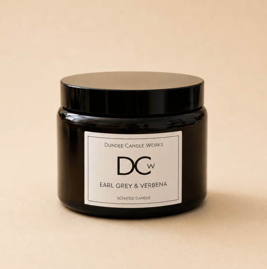 A dark amber glass jar candle with a black lid and a white label reading 'Dundee Candle Works DCw Earl Grey & Verbena Scented Candle' on a warm beige background.
