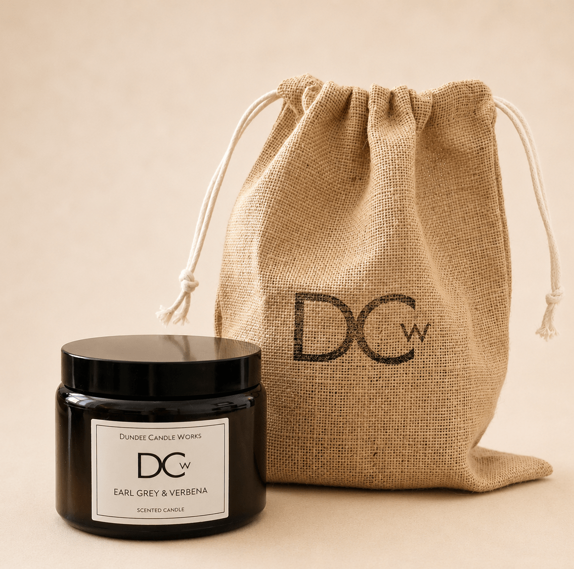 A black glass jar candle with a black lid and a white label reading 'Dundee Candle Works DCw Earl Grey & Verbena Scented Candle' sits in front of a natural jute drawstring pouch printed with 'DCw' on a warm beige background.