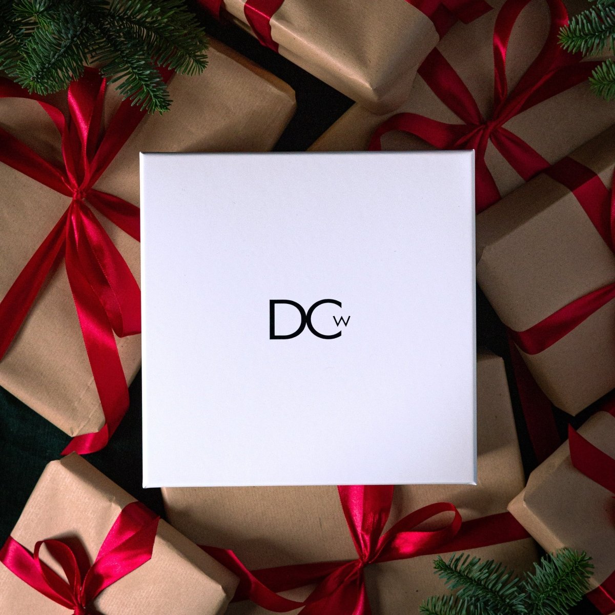 White gift box with 'DCW' logo surrounded by brown gift boxes with red ribbons and Christmas pine branches.