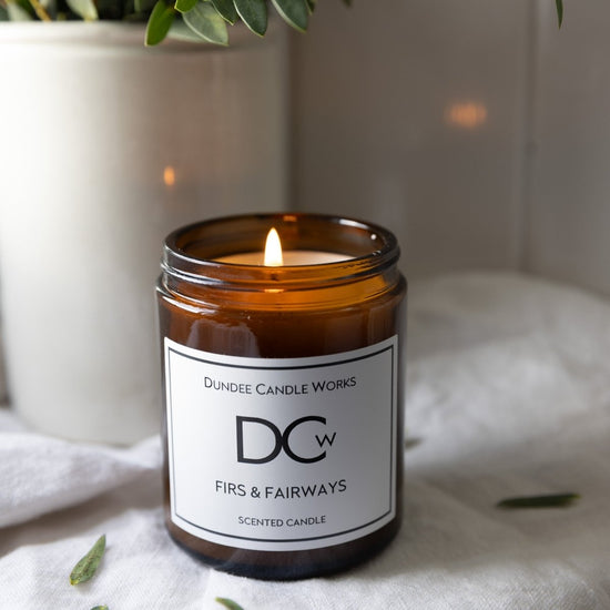 A lit amber glass jar candle with a white label reading 'Dundee Candle Works DCw Firs & Fairways Scented Candle,' placed on a white fabric surface with a softly blurred indoor background and greenery.