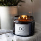 A lit three-wick scented candle in a dark amber glass jar with a white label reading 'Dundee Candle Works' and 'DCw Firs & Fairways', styled on a white fabric surface with a blurred potted plant in the background.