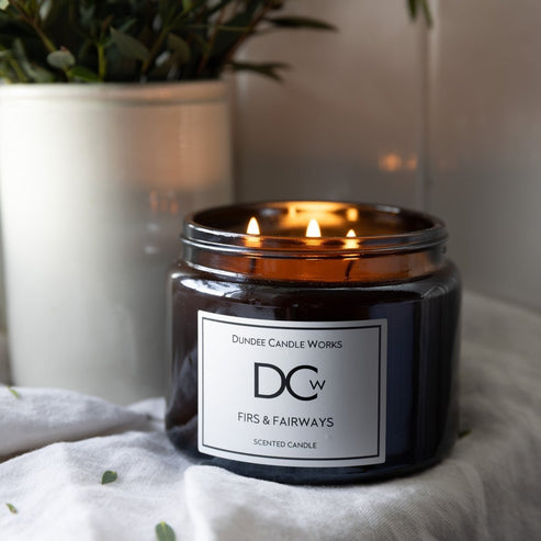 A lit three-wick scented candle in a dark amber glass jar with a white label reading 'Dundee Candle Works' and 'DCw Firs & Fairways', styled on a white fabric surface with a blurred potted plant in the background.