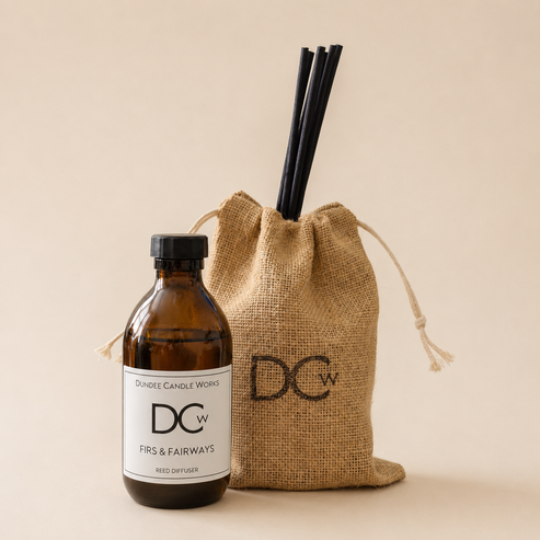 An amber glass reed diffuser bottle with a black cap and white label next to a natural jute drawstring bag with black reeds inserted, on a warm beige background. The label and bag both show the DCW logo and the scent name Firs & Fairways.