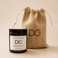 A scented candle in a dark amber glass jar with a black lid and a white label reading 'Dundee Candle Works DCw Firs & Fairways Scented Candle,' shown next to a natural jute drawstring pouch with a DCw logo, on a warm beige background.