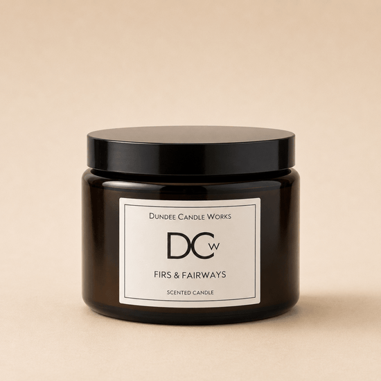 A dark amber glass jar candle with a black lid and a white label reading 'Dundee Candle Works DCw Firs & Fairways Scented Candle' on a warm beige background.