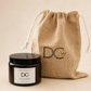 A black glass jar candle with a white label from Dundee Candle Works sits in front of a natural jute drawstring bag printed with the brand initials DCw, on a warm beige background.