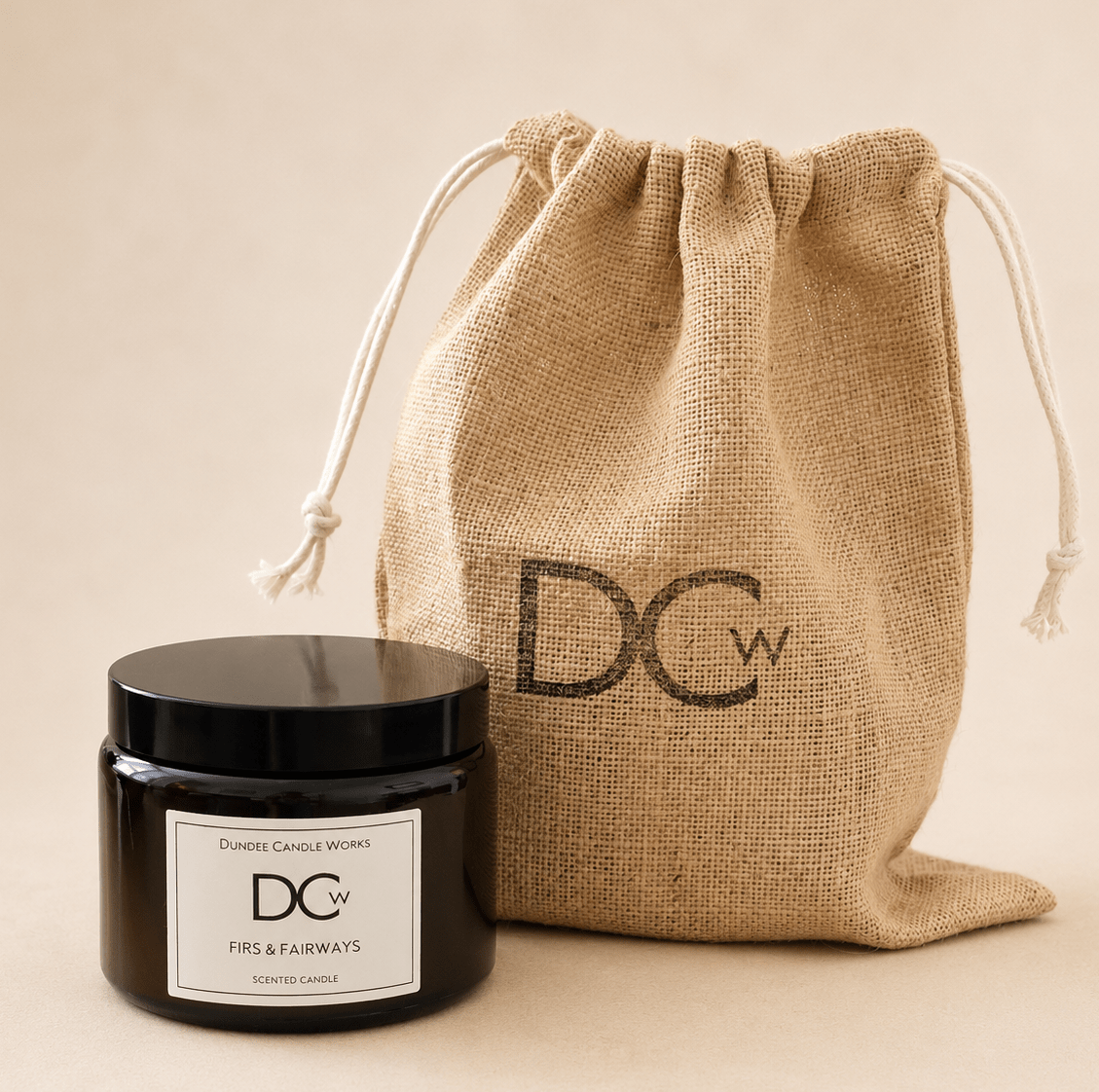 A black glass jar candle with a white label from Dundee Candle Works sits in front of a natural jute drawstring bag printed with the brand initials DCw, on a warm beige background.