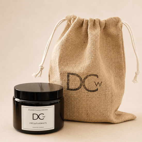 A black glass jar candle with a white label from Dundee Candle Works sits in front of a natural jute drawstring bag printed with the brand initials DCw, on a warm beige background.