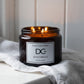A lit amber glass scented candle jar with three flames, labeled 'Dundee Candle Works Jute & Tobacco', resting on a white linen cloth against a soft white paneled background.