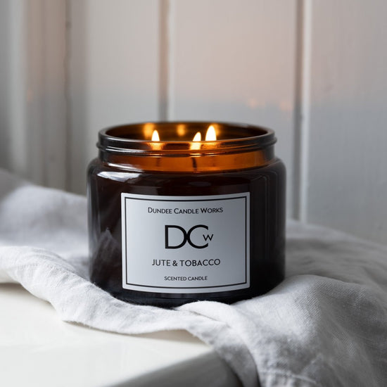 A lit amber glass scented candle jar with three flames, labeled 'Dundee Candle Works Jute & Tobacco', resting on a white linen cloth against a soft white paneled background.