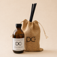 An amber glass bottle labeled Dancee Candle Works DC Jute & Tobacco Reed Diffuser stands next to a natural burlap drawstring bag with a black DCw logo and two black reed straws, on a warm beige background.