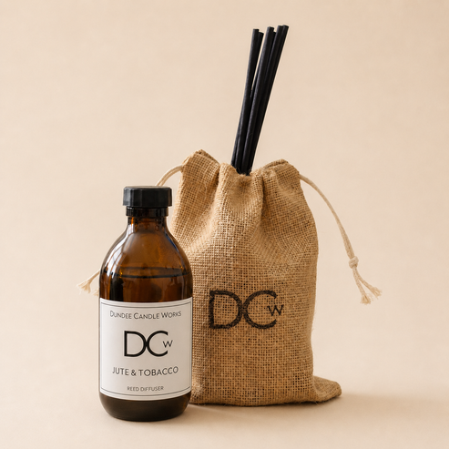 An amber glass bottle labeled Dancee Candle Works DC Jute & Tobacco Reed Diffuser stands next to a natural burlap drawstring bag with a black DCw logo and two black reed straws, on a warm beige background.