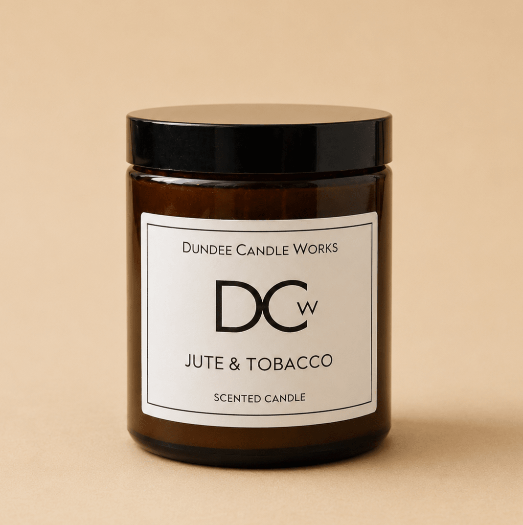 An amber glass jar candle with a black lid and a white label reading 'Dundee Candle Works DCw Jute & Tobacco Scented Candle' on a warm beige background.