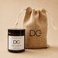 A Dundee Candle Works Jute & Tobacco scented candle in a dark amber glass jar with a black lid and white label, shown next to a natural jute drawstring pouch printed with the DCw logo. The background is a solid, warm beige tone.