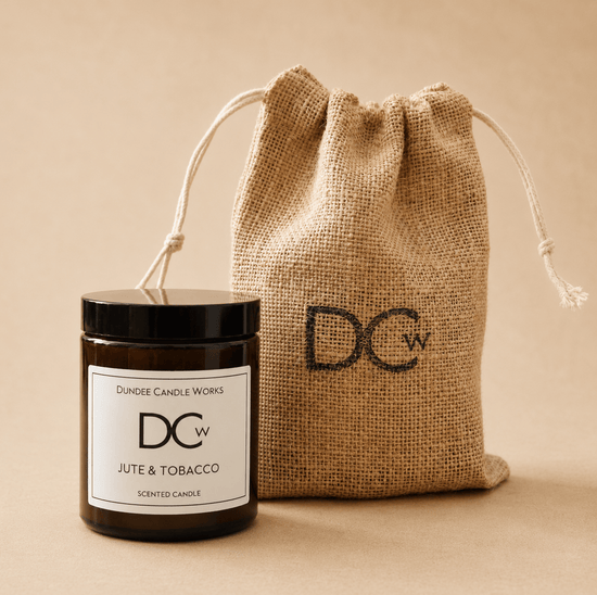 A Dundee Candle Works Jute & Tobacco scented candle in a dark amber glass jar with a black lid and white label, shown next to a natural jute drawstring pouch printed with the DCw logo. The background is a solid, warm beige tone.