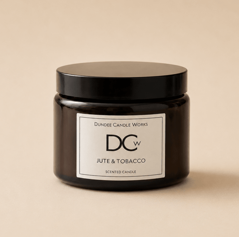 A dark amber glass jar candle with a black lid and a white label reading 'Dundee Candle Works DCw JUTE & TOBACCO SCENTED CANDLE' on a warm beige background.