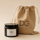 A Jute & Tobacco scented candle jar with a black lid and white label sits in front of a natural jute drawstring pouch. Both items display the DCW logo, set against a solid warm beige background.