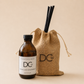 An amber glass Marmalade Gingerbread reed diffuser bottle with a black cap and white label next to a natural burlap drawstring bag with black reed diffuser straws, both branded DCW, on a warm beige background.