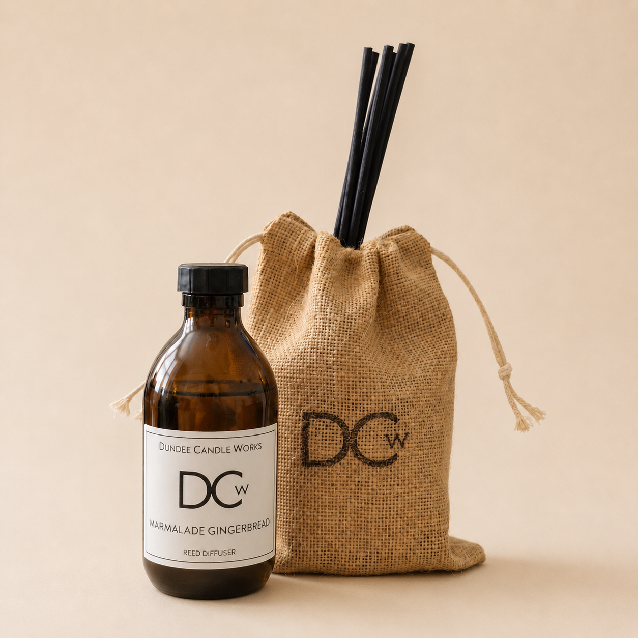 An amber glass Marmalade Gingerbread reed diffuser bottle with a black cap and white label next to a natural burlap drawstring bag with black reed diffuser straws, both branded DCW, on a warm beige background.