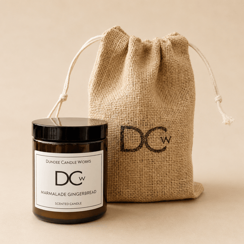 A Dundee Candle Works single wick Marmalade GIngerbread scented candle in a dark amber glass jar with a black lid and white label, standing next to a natural burlap drawstring pouch printed with the DCw logo. The background is a solid, warm beige studio backdrop.