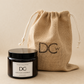 A DCW Marmalade Gingerbread scented candle in a black glass jar with a black lid and white label, displayed next to its natural burlap drawstring pouch which features a black DCW logo, on a neutral beige background.
