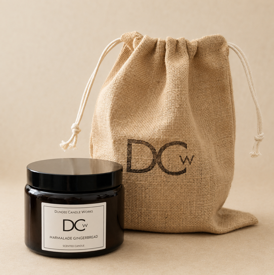 A DCW Marmalade Gingerbread scented candle in a black glass jar with a black lid and white label, displayed next to its natural burlap drawstring pouch which features a black DCW logo, on a neutral beige background.