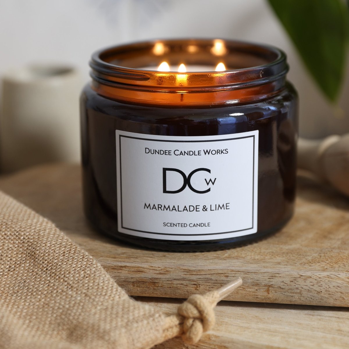 A lit amber glass jar candle from Dundee Candle Works, labeled 'Marmalade & Lime', resting on a wooden surface with a jute bag nearby.