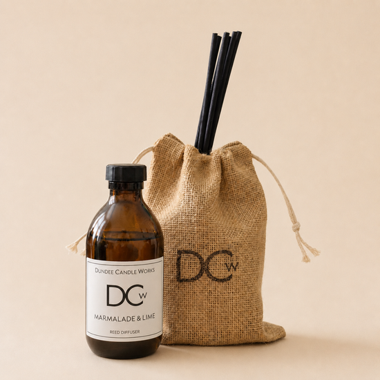 An amber glass reed diffuser bottle with a black cap and white label stands beside a natural jute drawstring pouch containing black reed sticks. The label reads Dundee Candle Works and the scent is Marmalade & Lime.