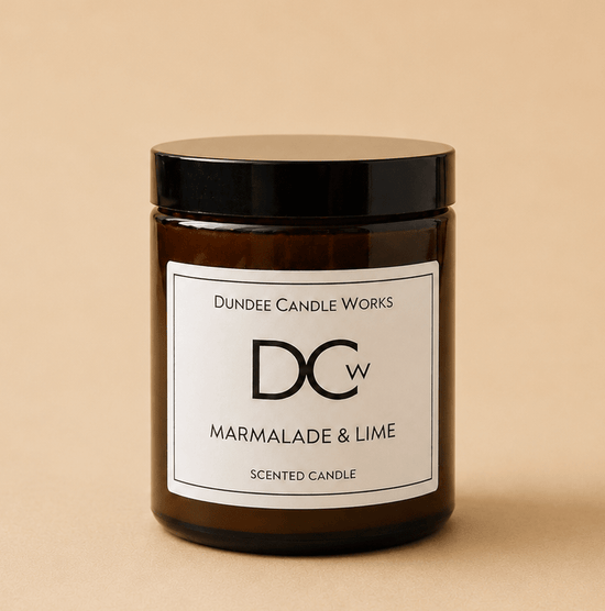 An amber glass jar candle with a black lid and a white label reading 'Dundee Candle Works DCw Marmalade & Lime Scented Candle' on a warm beige background.