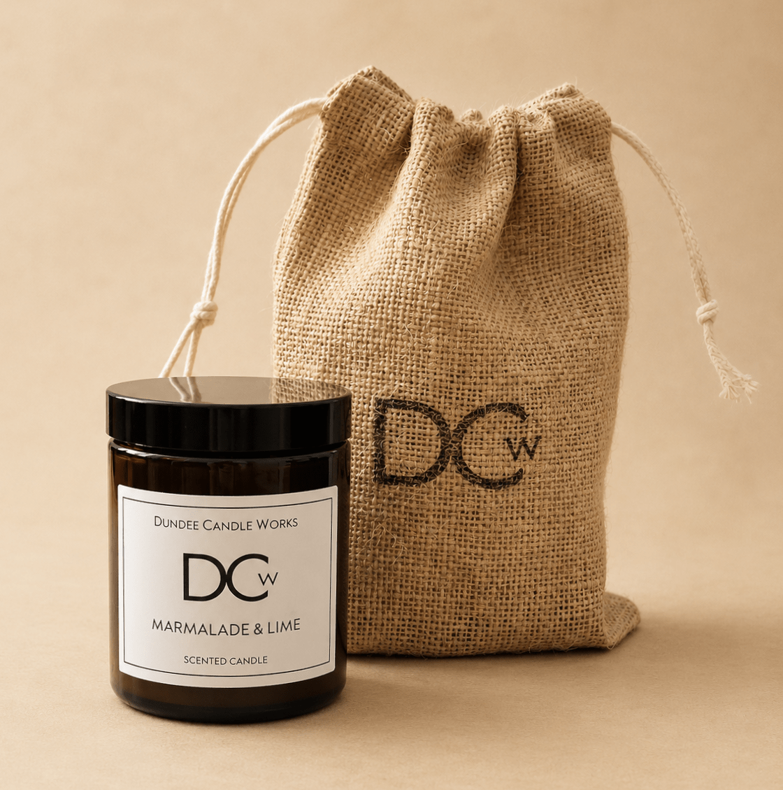 A scented candle jar with a black lid and white label in front of a natural jute drawstring pouch. The label reads 'Dundee Candle Works' and 'Marmalade & Lime', and the candle and pouch both have a 'DCw' logo.