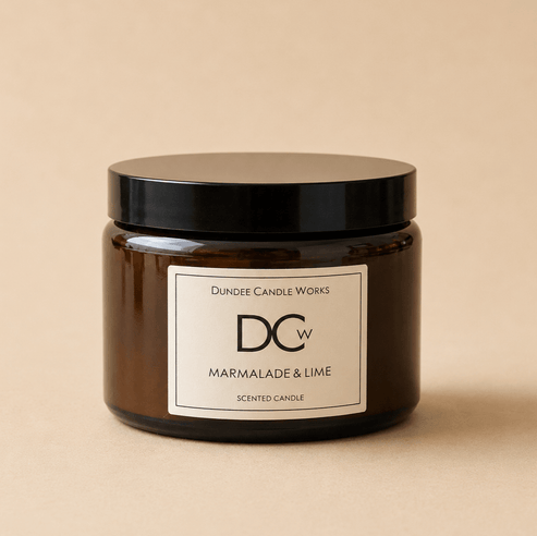 A dark amber glass jar candle with a black lid and a white label reading 'Dundee Candle Works DCw Marmalade & Lime Scented Candle' on a warm beige background.